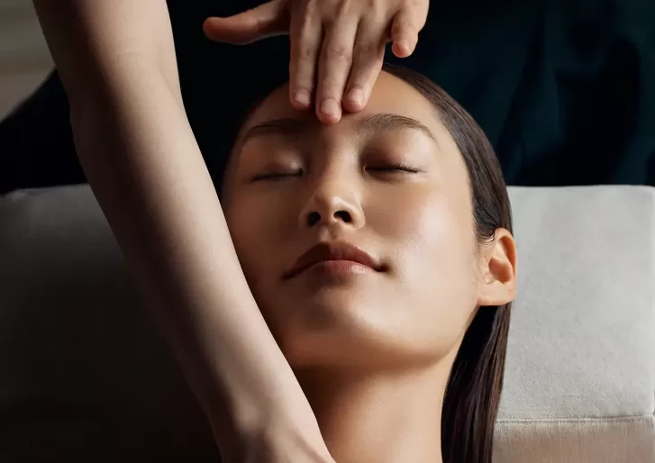 A person with closed eyes receiving a facial massage. Two hands gently press on the persons forehead and cheek. The setting is calm and relaxing, suggesting a spa or wellness environment at La Mer at the Ritz Carlton Hong Kong.