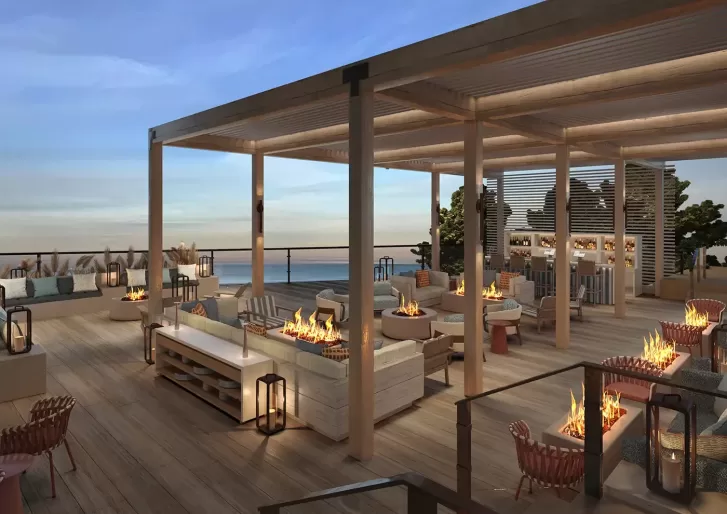 Rooftop lounge at sunset with modern seating, fire pits, wooden pergolas, and ocean views. Cozy sofas and chairs are arranged around low tables, creating an inviting atmosphere for relaxation.