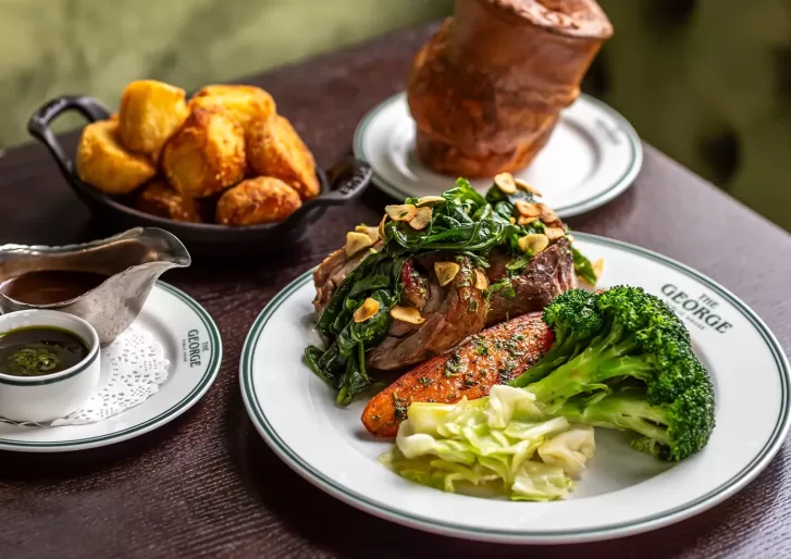A plate with roast beef topped with greens and garlic, served with carrots, broccoli, and cabbage. Nearby are a dish of sauce, a bowl of golden roast potatoes, and a large Yorkshire pudding.