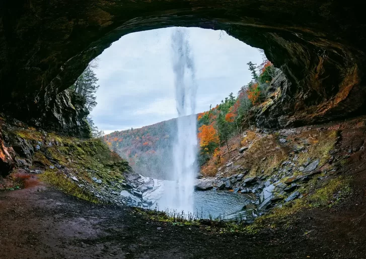 View from behind a waterfall cascading from a cave opening, with a serene pool below. The surrounding landscape is lush with autumn foliage in hues of orange and red, under an overcast sky.