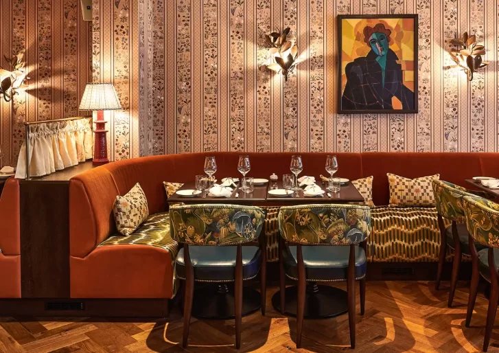 Cosy restaurant interior featuring a curved orange booth with patterned pillows, surrounded by green, floral-upholstered chairs, a patterned wallpaper, and a colorful abstract painting. Tables are set with glasses and napkins.
