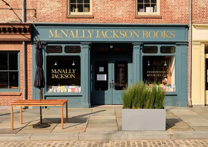 Exterior of McNally Jackson Books in NYC with a green facade and gold lettering. The storefront features large windows displaying books, a small table, a closed umbrella, and a planter with tall grasses on the cobblestone sidewalk.