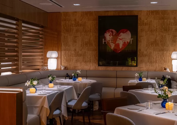 A cozy, dimly lit restaurant with white tablecloths, modern chairs, floral centerpieces, and glowing candles. A large heart artwork with the word HEART hangs on the wall behind a curved booth.