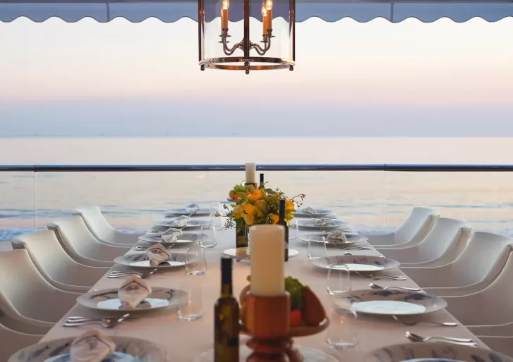 A long dining table set for a meal with white chairs, plates, glasses, and floral centerpieces sits on a terrace overlooking the calm sea at sunset. A chandelier hangs above the table.