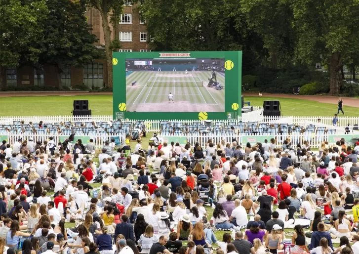 A large crowd sits on grass watching a live tennis match on a giant outdoor screen in a park, surrounded by trees and buildings. The atmosphere is lively and festive.