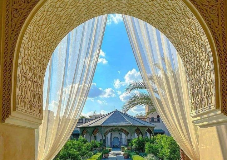View through an ornate arched doorway with sheer curtains revealing a lush garden, palm trees, and a pavilion under a bright blue sky with scattered clouds. Intricate patterns decorate the arch.