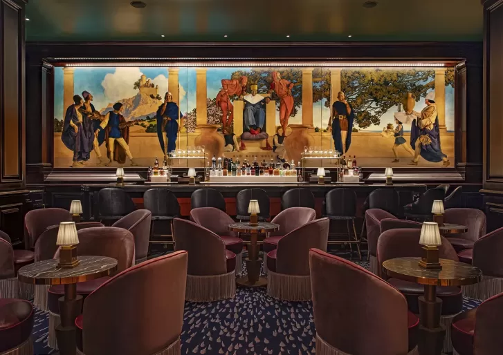 A dimly lit bar with plush velvet chairs and small tables equipped with lamps. A large, colorful mural depicting a historical scene is behind the well-stocked bar counter, adding an artistic touch to the sophisticated setting.