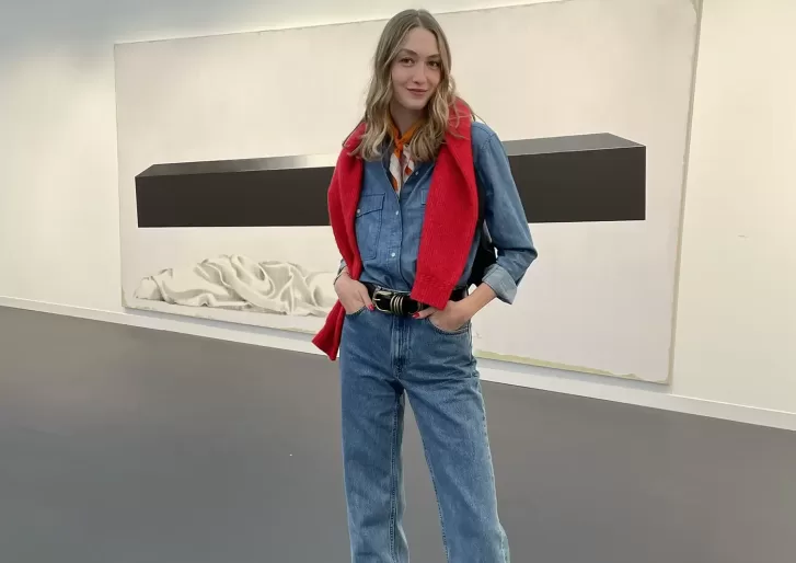 A woman stands indoors wearing a denim shirt and jeans, with a red sweater draped over her shoulders. She poses with one hand in her pocket, smiling slightly. Behind her is a large, minimalist artwork on the wall.