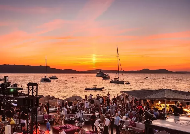 A vibrant beach club scene at sunset, with many people socializing near the shore. Boats float on the calm sea, and colorful clouds fill the sky as the sun sets in the background.