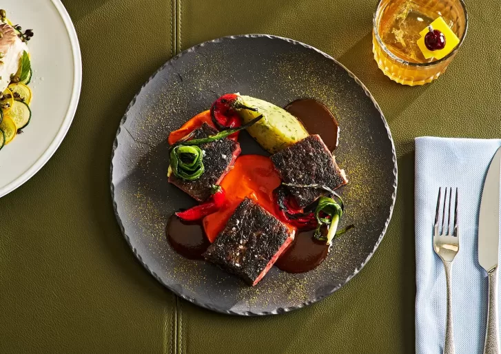 A dish with seared meat, drizzled with red and brown sauce, garnished with roasted red peppers and greens on a black plate. An avocado slice is at the top. Beside it, a glass with an orange liquid and ice, garnished with a cherry, is visible.