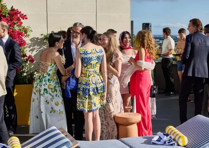 A group of people dressed in colorful, semi-formal summer attire socialize on a sunny rooftop patio. Flowers, drinks, and city views are visible, creating a lively and festive atmosphere.