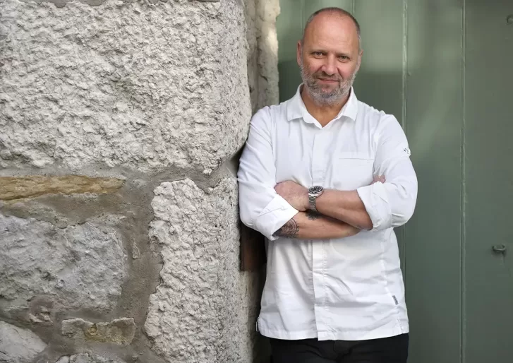 Chef Simon Rogan, wearing a white chefs coat, stands with arms crossed against a stone wall next to a green door, smiling slightly at the camera.