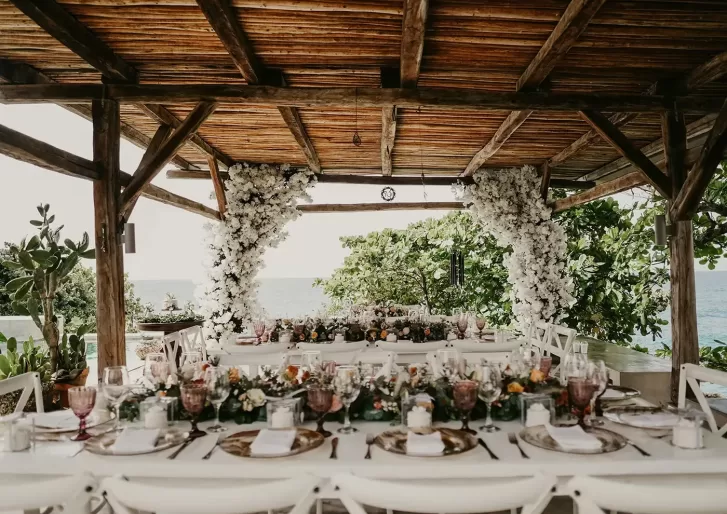 A decorated outdoor dining area under a wooden pergola, featuring long tables set with elegant tableware, floral centerpieces, and white flowers arching around the structure, with greenery and the ocean visible in the background.