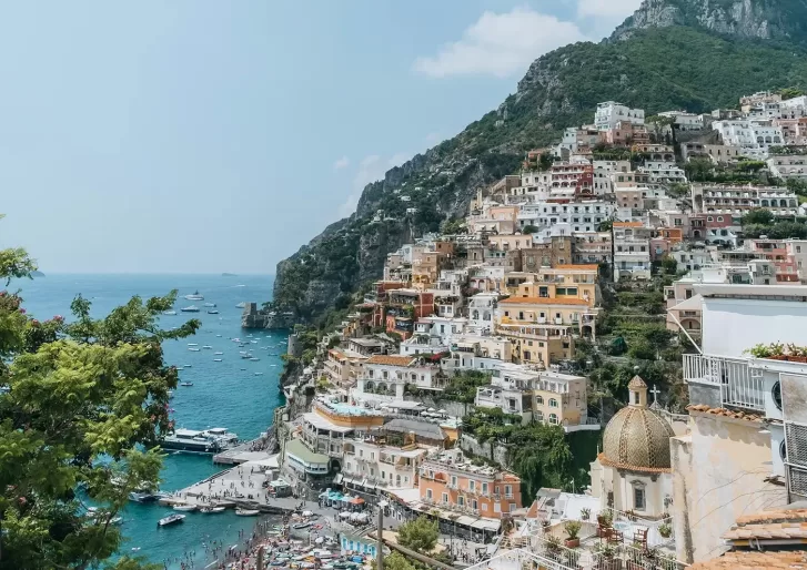 Colorful buildings stacked on a steep hillside overlooking a sparkling blue sea with boats, lush greenery, and a crowded beach below; mountains rise in the background under a clear, sunny sky.