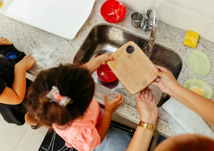 A child and an adult are washing a small wooden cutting board in a kitchen sink. The countertop is cluttered with soap, sponges, and a red bowl. The child is wearing a pink shirt, and the adult is wearing a watch on the left wrist.