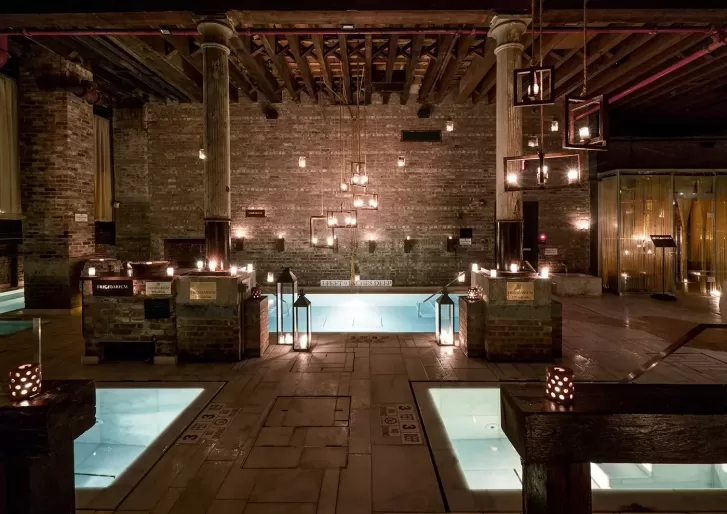 Spa interior with dim lighting features several rectangular pools surrounded by rustic stone walls and wooden beams. Candles and hanging lanterns illuminate the serene space. Steps lead into the central pool.