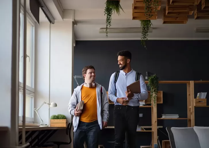 Two people walking and smiling in a modern office space with shelves and plants. One holds a laptop and a tablet, while the other carries books. Bright window light illuminates the room.