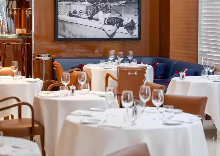 Elegant restaurant interior with round tables covered in white tablecloths, set with glassware and cutlery. Brown leather chairs surround the tables. A black and white photo hangs on a wood-paneled wall in the background.