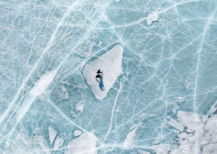 A person stands on a white patch of ice, surrounded by blue cracked and textured frozen lake surface, casting a long shadow. The scene is viewed from above.