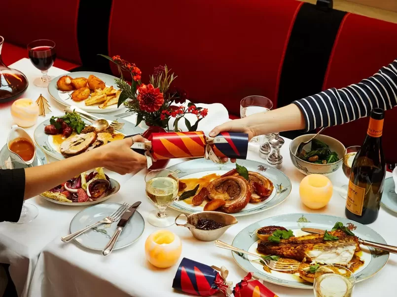 A festive dinner table with two people pulling a Christmas cracker. Plates feature roasted meat, vegetables, and a side dish. The table is decorated with candles, flowers, and a glass of white wine at Ham Yard Hotel, Soho in London.