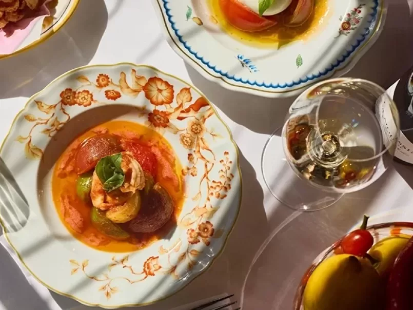 A sunlit table set with ornate plates holding colorful tomato salads and a glass of white wine, surrounded by decorative cutlery and a bowl of fresh tomatoes.