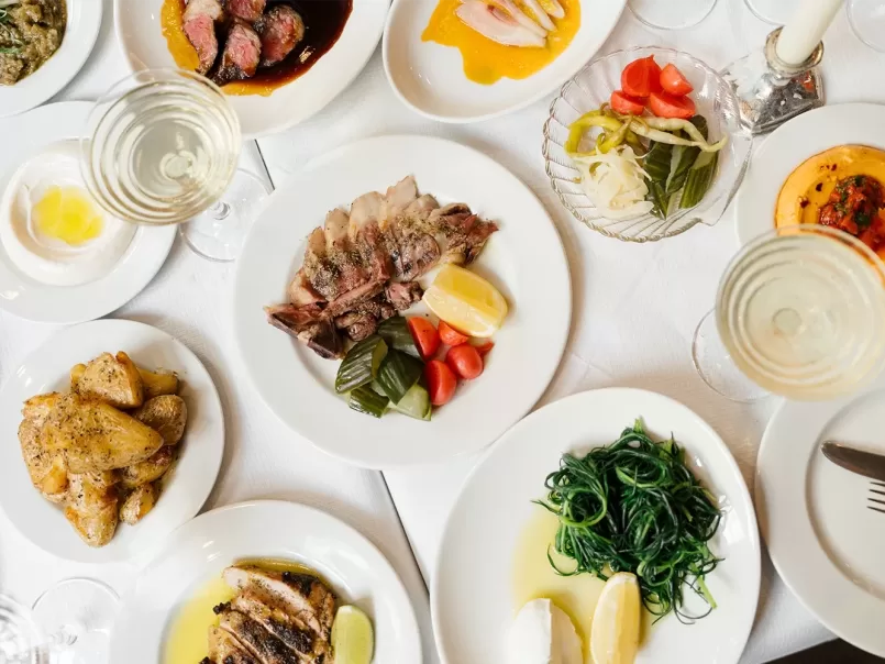 A table set with various plated dishes, including sliced meat with lemon and vegetables, sautéed greens, roasted potatoes, pickled vegetables, and glasses of white wine on a white tablecloth.
