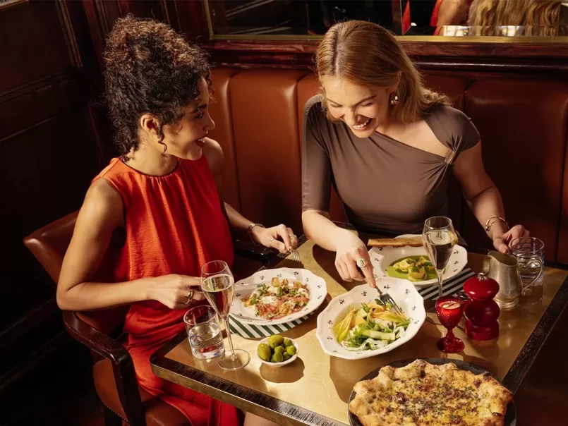 Two women sit at a restaurant table enjoying pasta dishes, salad, and pizza. They are smiling and conversing, with glasses of water and wine in front of them on a cozy brown leather booth.