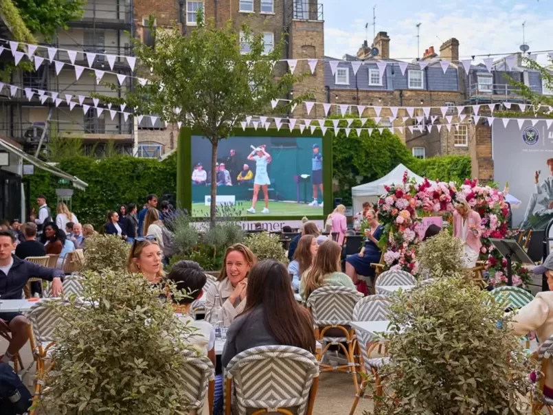 People sit at outdoor tables in a garden setting, watching a large screen showing a tennis match. The area is decorated with bunting and flowers, creating a festive, social atmosphere. Buildings and greenery are in the background.