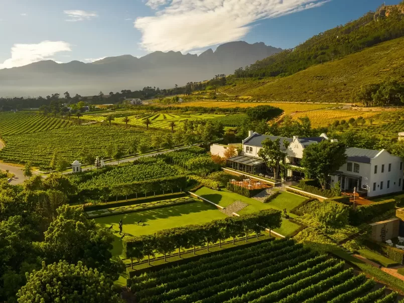 Aerial view of a lush vineyard estate with a white house, manicured gardens, and rows of grapevines, surrounded by green hills and distant mountains under a partly cloudy sky.