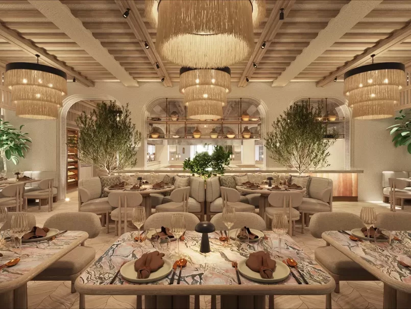 Elegant restaurant interior with soft lighting, large fringe chandeliers, cozy beige seating, marble-topped tables set with glassware and napkins, lush green plants, and an open kitchen in the background.
