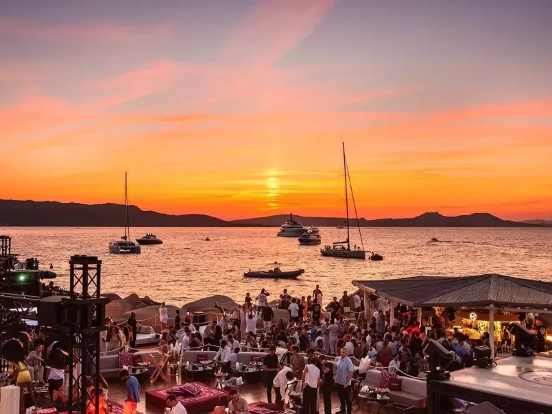 A vibrant beach club scene at sunset, with many people socializing near the shore. Boats float on the calm sea, and colorful clouds fill the sky as the sun sets in the background.
