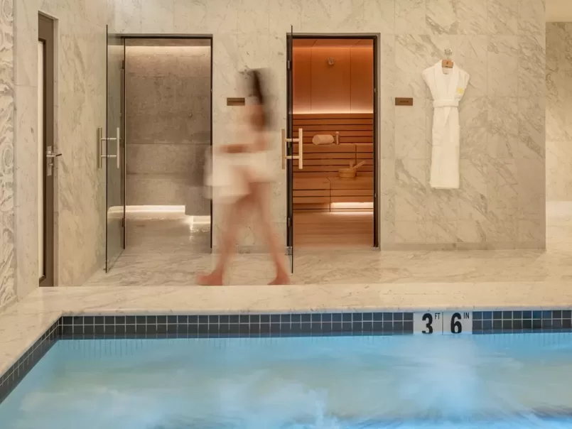 A blurred person in a towel walks past a sauna and steam room in a marble spa area, with a robe hanging on the wall and a pool with bubbling water in the foreground marked 3 ft 6 in.