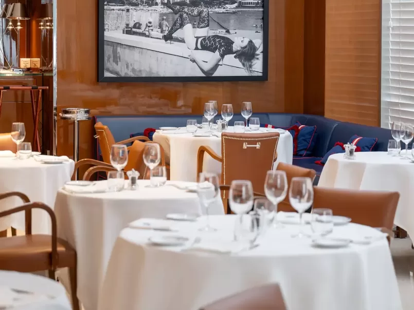 Elegant restaurant interior with round tables covered in white tablecloths, set with glassware and cutlery. Brown leather chairs surround the tables. A black and white photo hangs on a wood-paneled wall in the background.