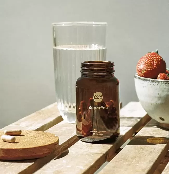 Table with bottle of Moon Juice SuperYou, glass of water, bowl of strawberries and two vitamins on a coaster