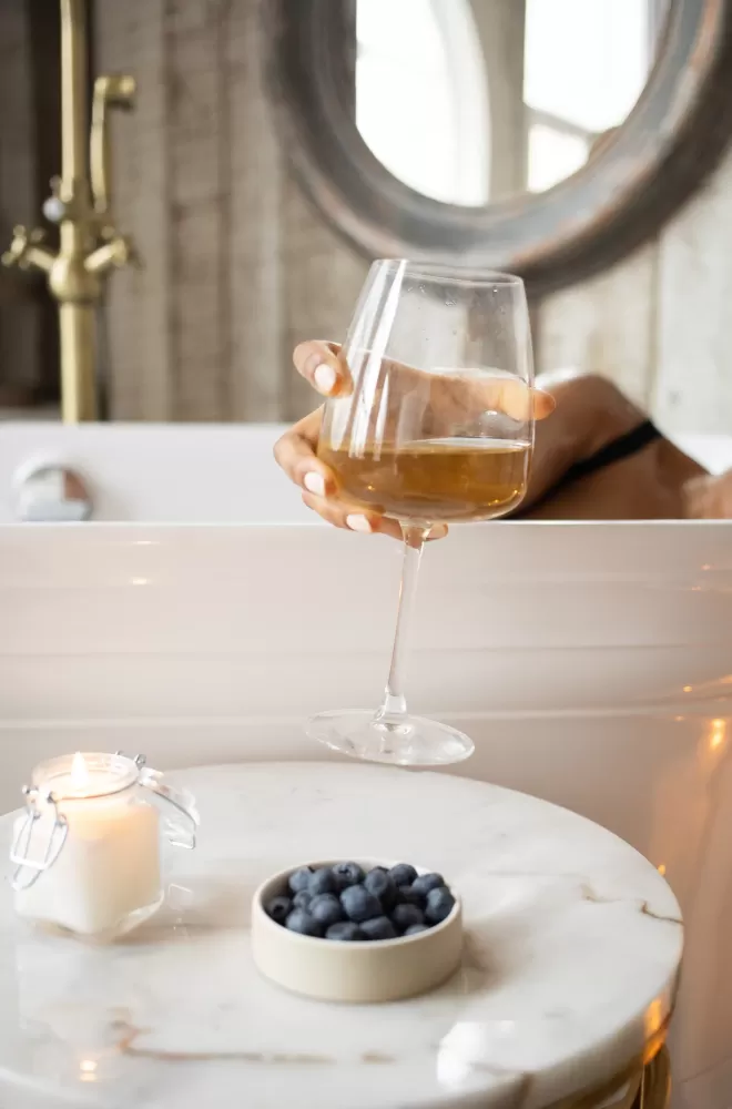 Women in bath reaching for glass of wine from table with bluebrries