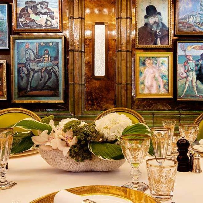 Grand dining table set up with flowers and abstract wine glasses facing a wall on portrait paintings on a wooden wall