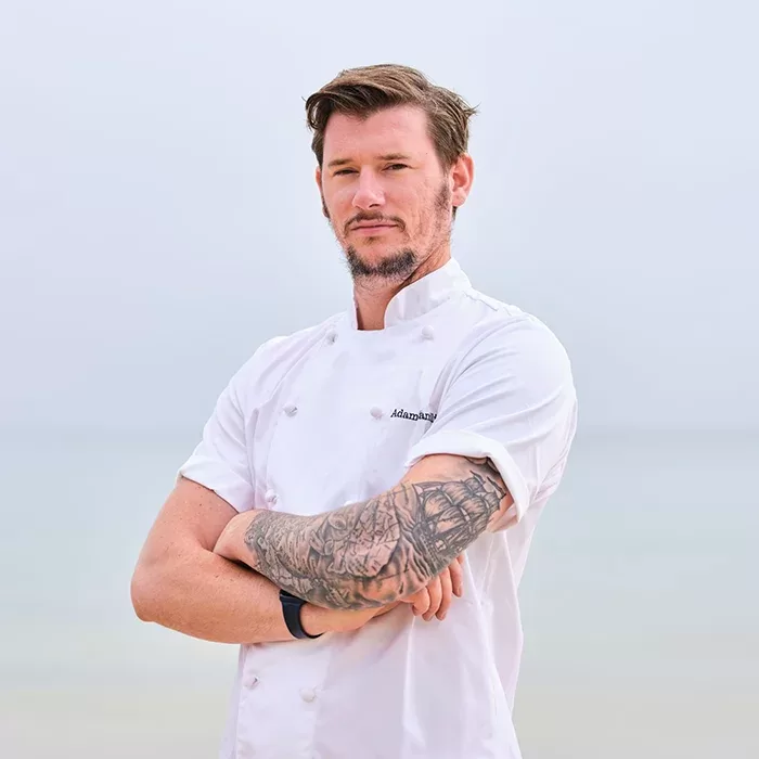 A man with short brown hair and a beard, wearing a white chefs coat with folded arms, stands outdoors against a blurred sky and water background. He has a large tattoo on his right forearm.