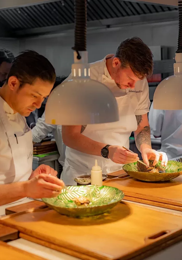 Two chefs in white uniforms carefully plate food under bright pendant lights in a professional kitchen, focusing on presentation with large green glass dishes in front of them.