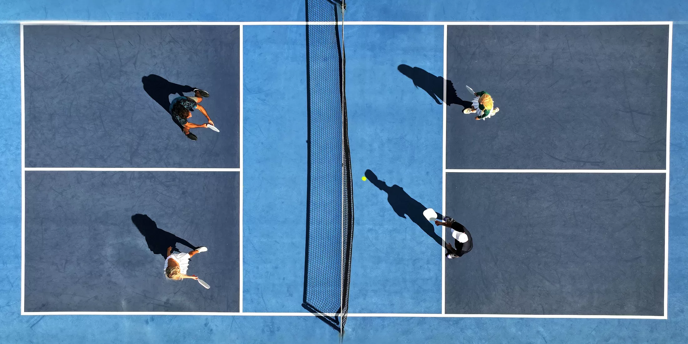 Aerial view of four people playing doubles pickleball on a blue court, with two players on each side of the net and their shadows visible on the court.
