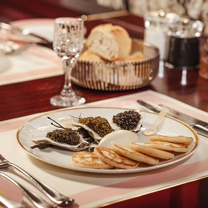 A plate with oysters topped with caviar, a serving of sour cream, mini pancakes (blinis), and a mother-of-pearl spoon on a set dining table; bread and a wine glass are in the background.