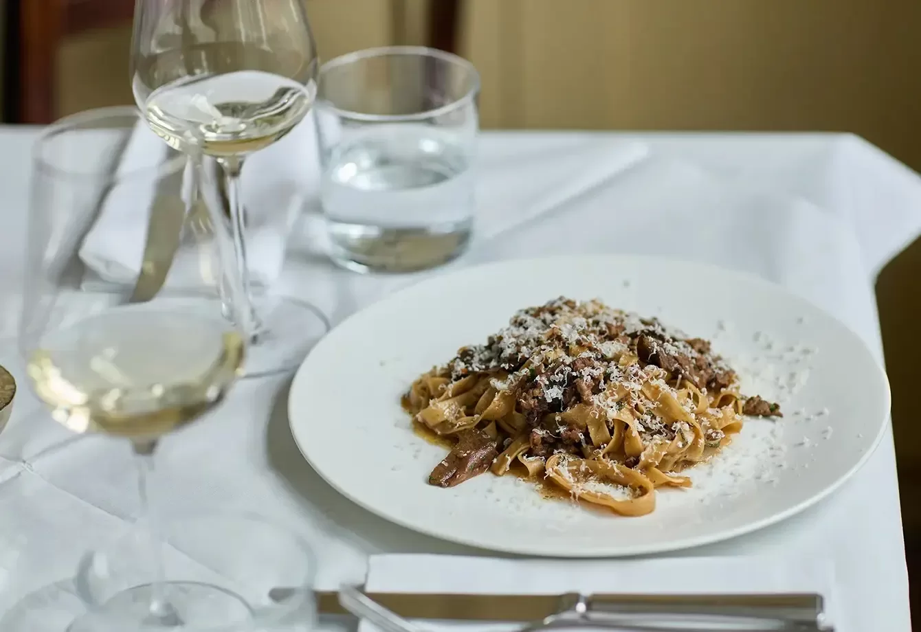 A plate of pasta topped with grated cheese and meat sauce on a white tablecloth, accompanied by a glass of white wine, a water glass, and cutlery.