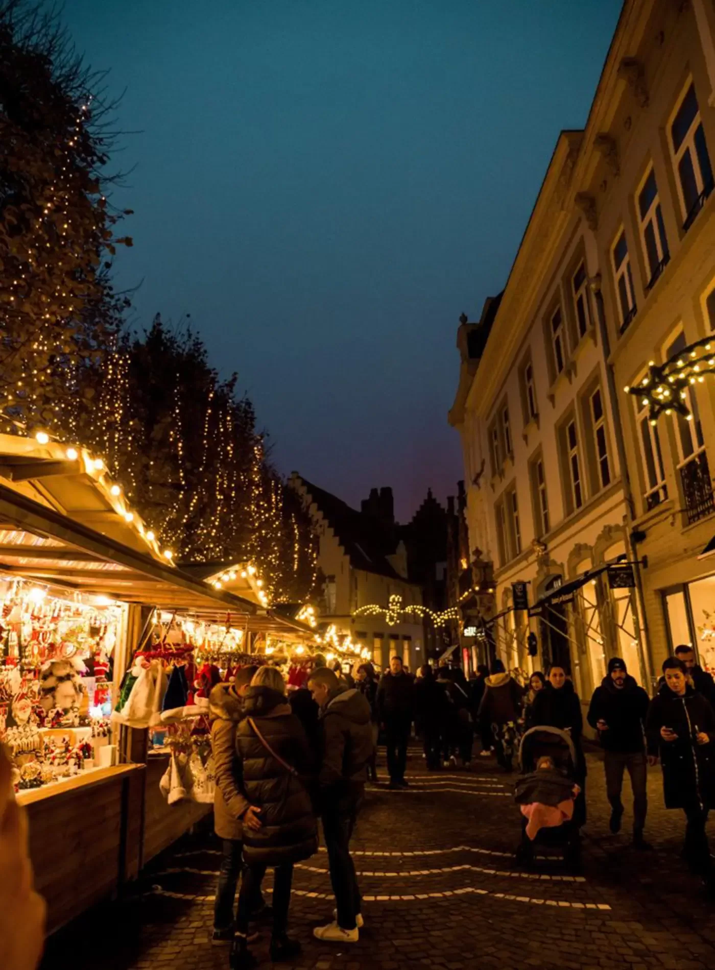 A festive outdoor market at dusk with people browsing decorated stalls and twinkling string lights hanging from trees and buildings; the atmosphere is lively and warm.
