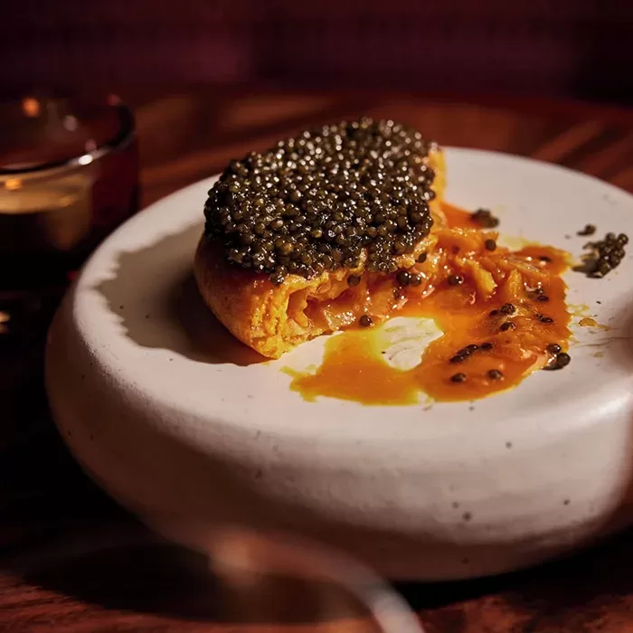 A thick piece of bread topped with a generous layer of black caviar sits on a white plate, with a rich orange sauce oozing out from underneath. A glass is partially visible in the background.