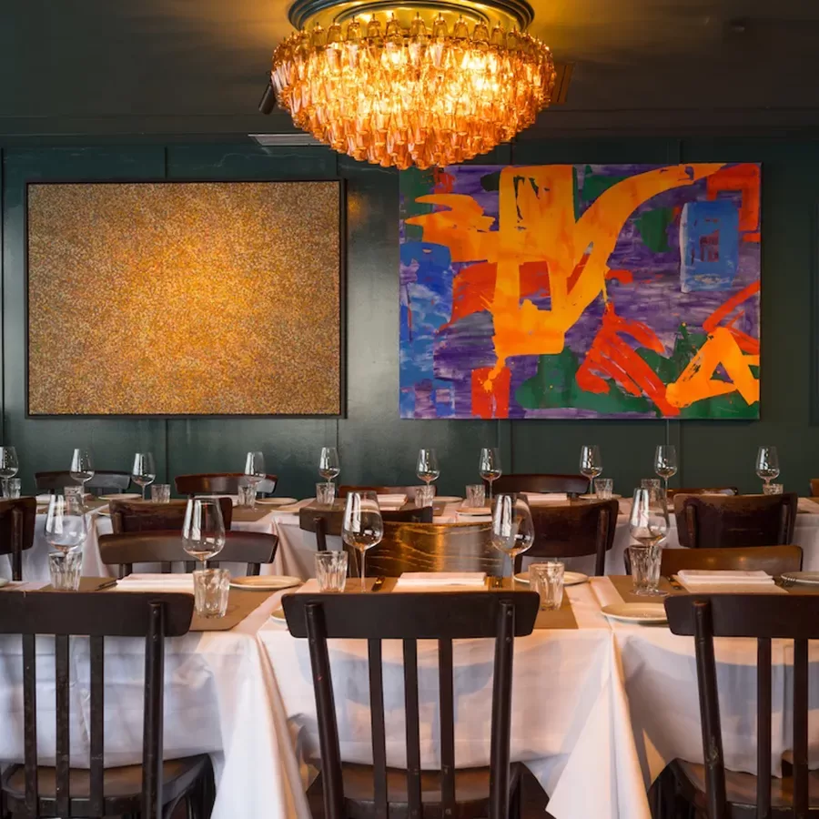A stylish restaurant dining area with tables covered in white cloths, set with wine glasses and plates, dark wooden chairs, a gold chandelier, and colorful abstract artwork on the green walls.