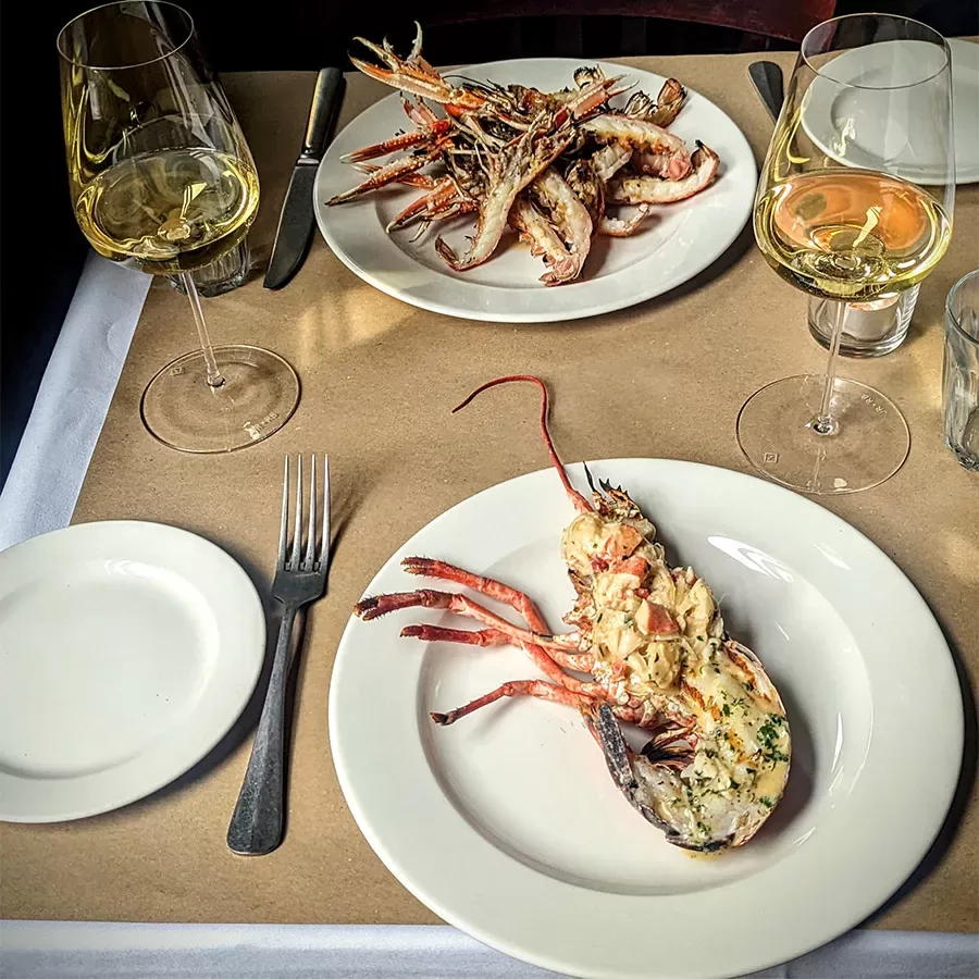 Two plates of lobster, each with half a lobster garnished with herbs, are set on a table beside two glasses of white wine, a fork, knife, empty plate, and a glass of water.