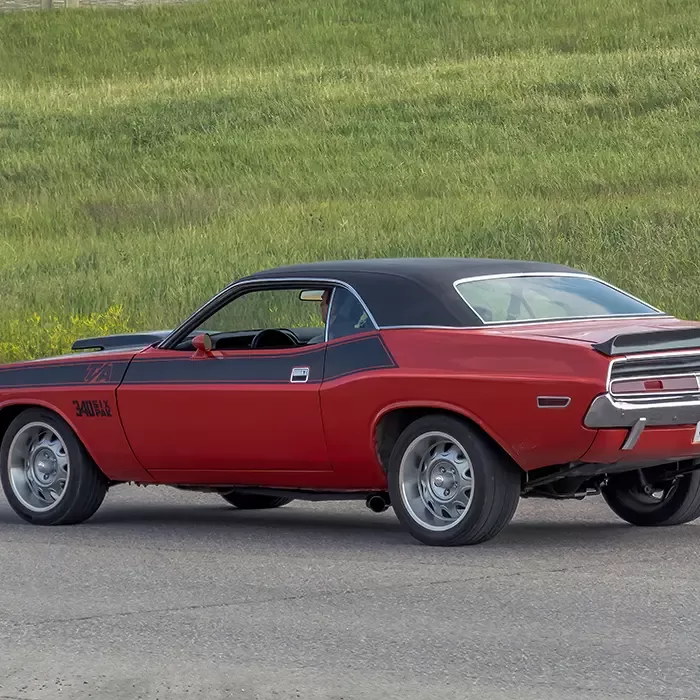A classic red muscle car with a black roof and rear spoiler is parked on a paved road, with green grass and a hillside in the background.