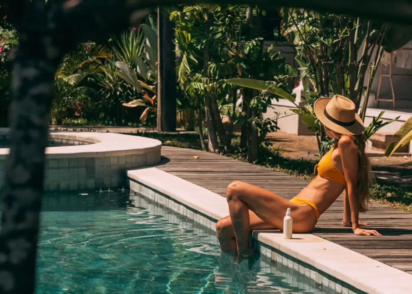 A woman in a yellow bikini and straw hat sits at the edge of a pool with her feet in the water, surrounded by lush tropical plants, with a bottle placed beside her on the poolside.