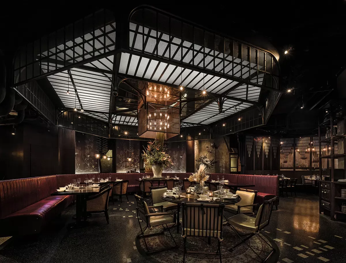 A luxurious, dimly lit restaurant interior with dark tones, featuring plush burgundy seating, ornate lighting fixtures, and an elegant centerpiece of floral arrangements. The space exudes a sophisticated and intimate ambiance at Mott 32, Hong Kong.