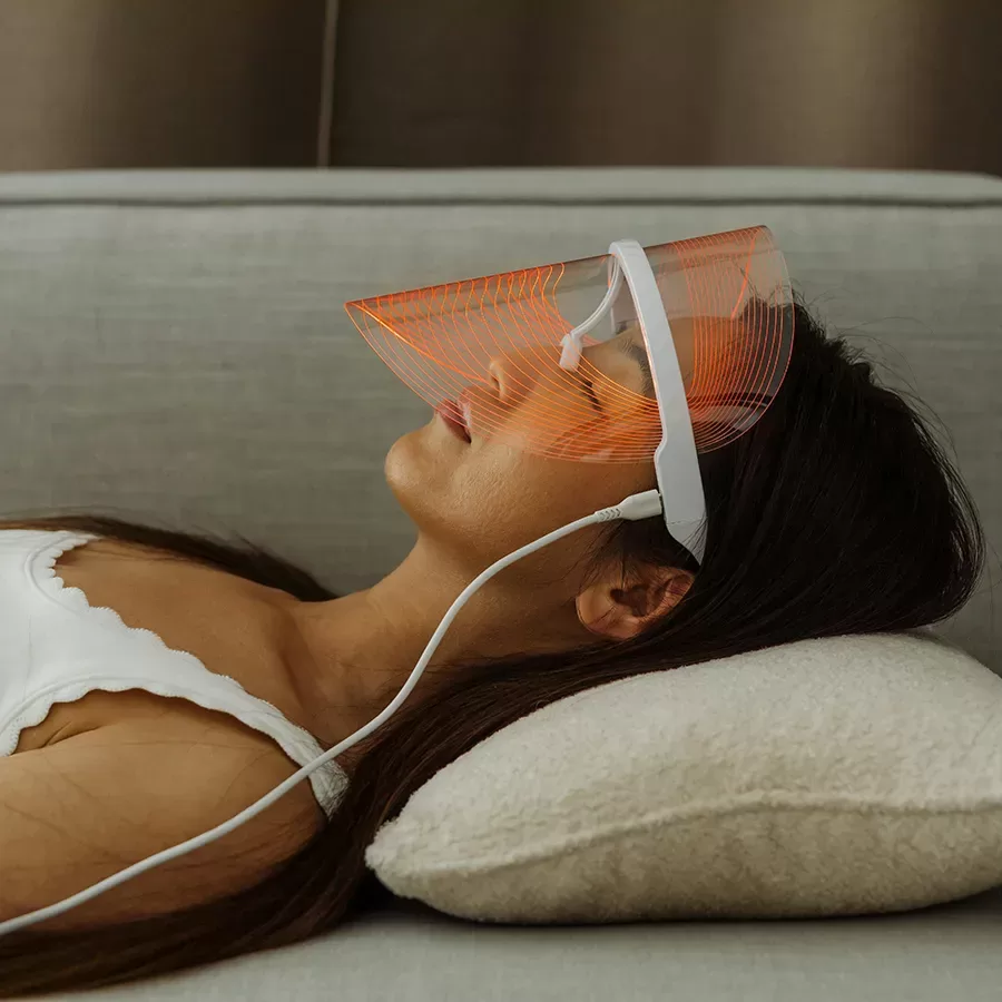 A woman lies on a pillow wearing a futuristic LED light therapy mask with orange lights, connected by a white cord, while resting on a gray sofa.