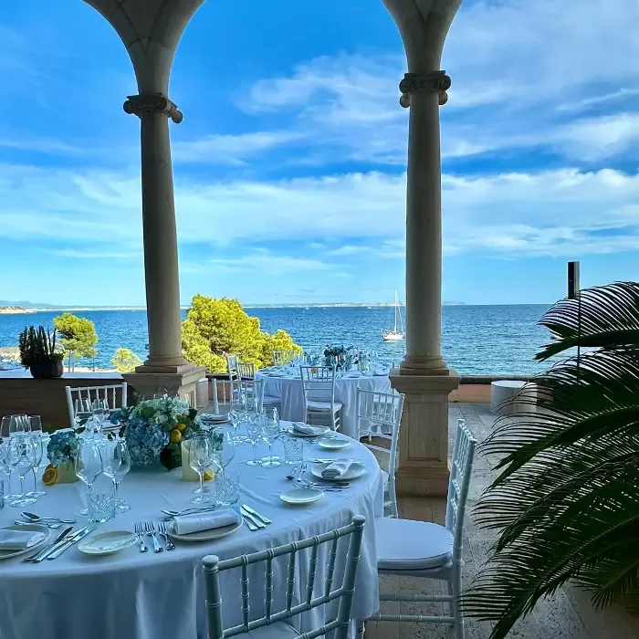 Elegant outdoor dining setup with round tables and white chairs on a terrace overlooking the sea, framed by stone columns. Blue sky, scattered clouds, sailboat, and greenery enhance the scenic, tranquil atmosphere.